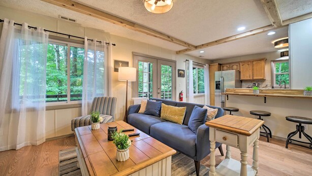 A cozy living room with a sofa, coffee table, and a small wooden side table. There are large windows with sheer curtains, a ceiling fan, and a kitchen area with wooden cabinets and a countertop.