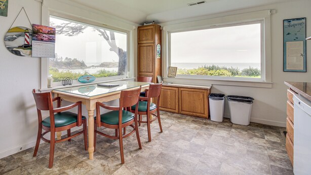 Una cocina con mesa y sillas, ventanas amplias con vista al océano, un armario de madera y dos cubos de basura.