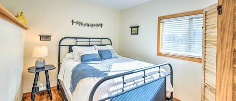 A neatly arranged bedroom with a metal bed frame, a bedside table with a lamp, a window with blinds, and a small shelf with decorative items.