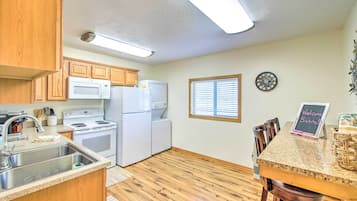 A kitchen with wooden cabinets, a microwave, a stove, and a refrigerator. There is a window with blinds, a clock on the wall, and a welcome sign on the counter.