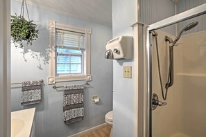 A bathroom with a toilet, a sink, a shower with a handheld showerhead, a towel rack with patterned towels, a window with blinds, and a wall-mounted heater. - Bay St. Louis House w/ Boat Dock & Launch! (Bay St Louis)