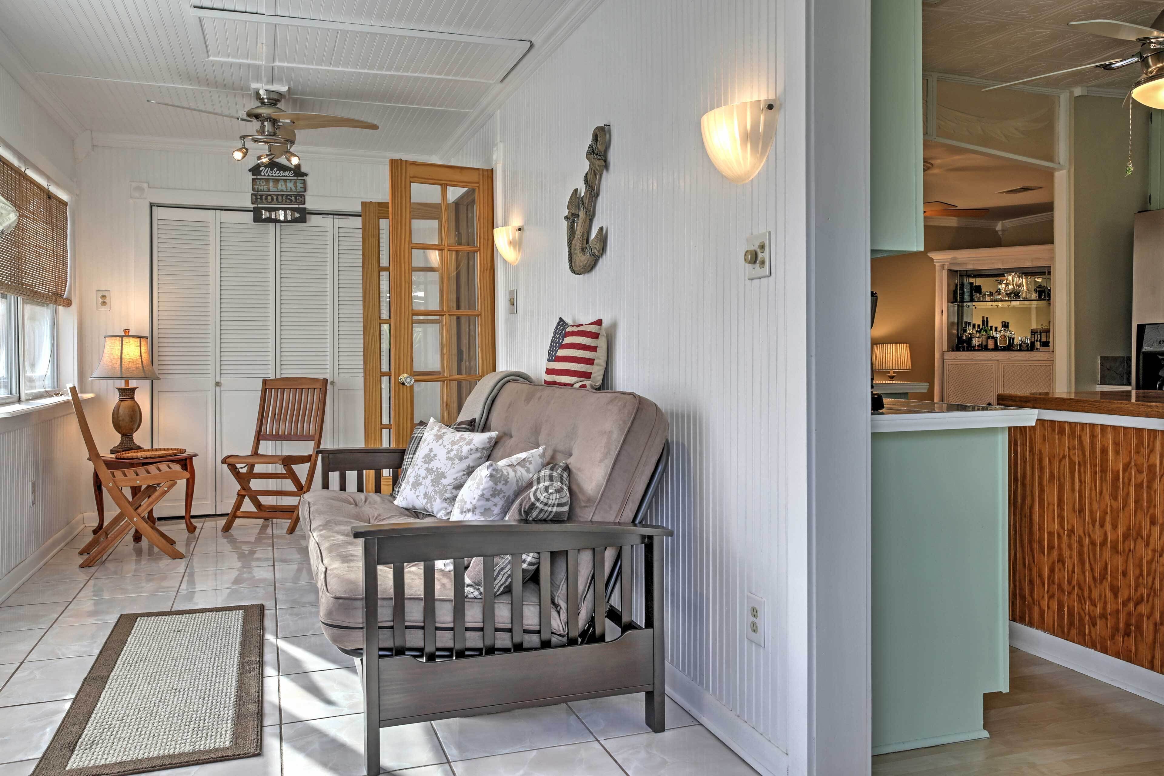 Un salon chaleureux avec un canapé gris, des chaises en bois, une petite table et un berceau. On aperçoit à l’arrière-plan un coin cuisine avec un bar.