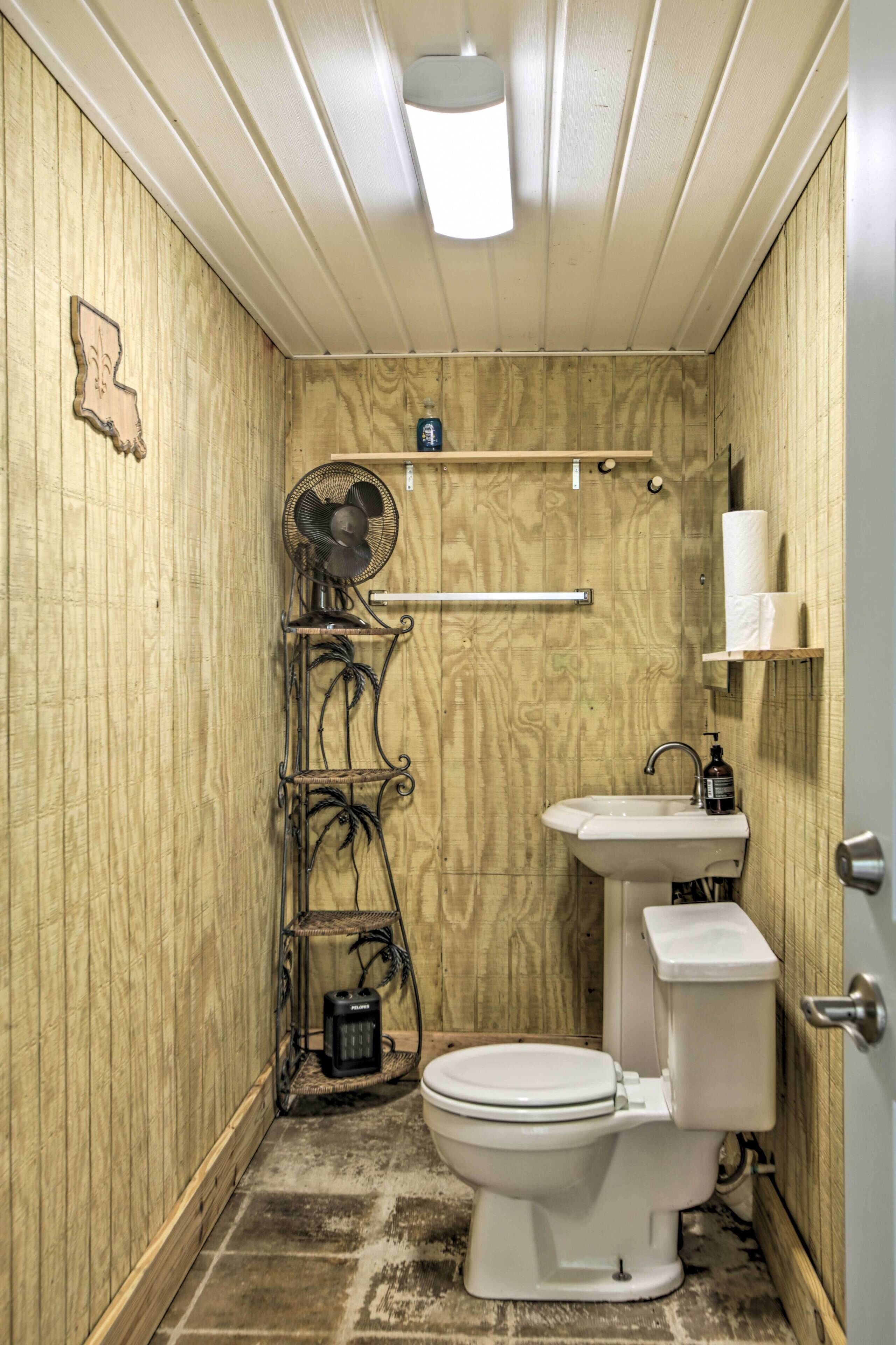 Une petite salle de bain compacte avec un mur en bois, des toilettes blanches, un lavabo, un ventilateur et une étagère contenant des articles de toilette.