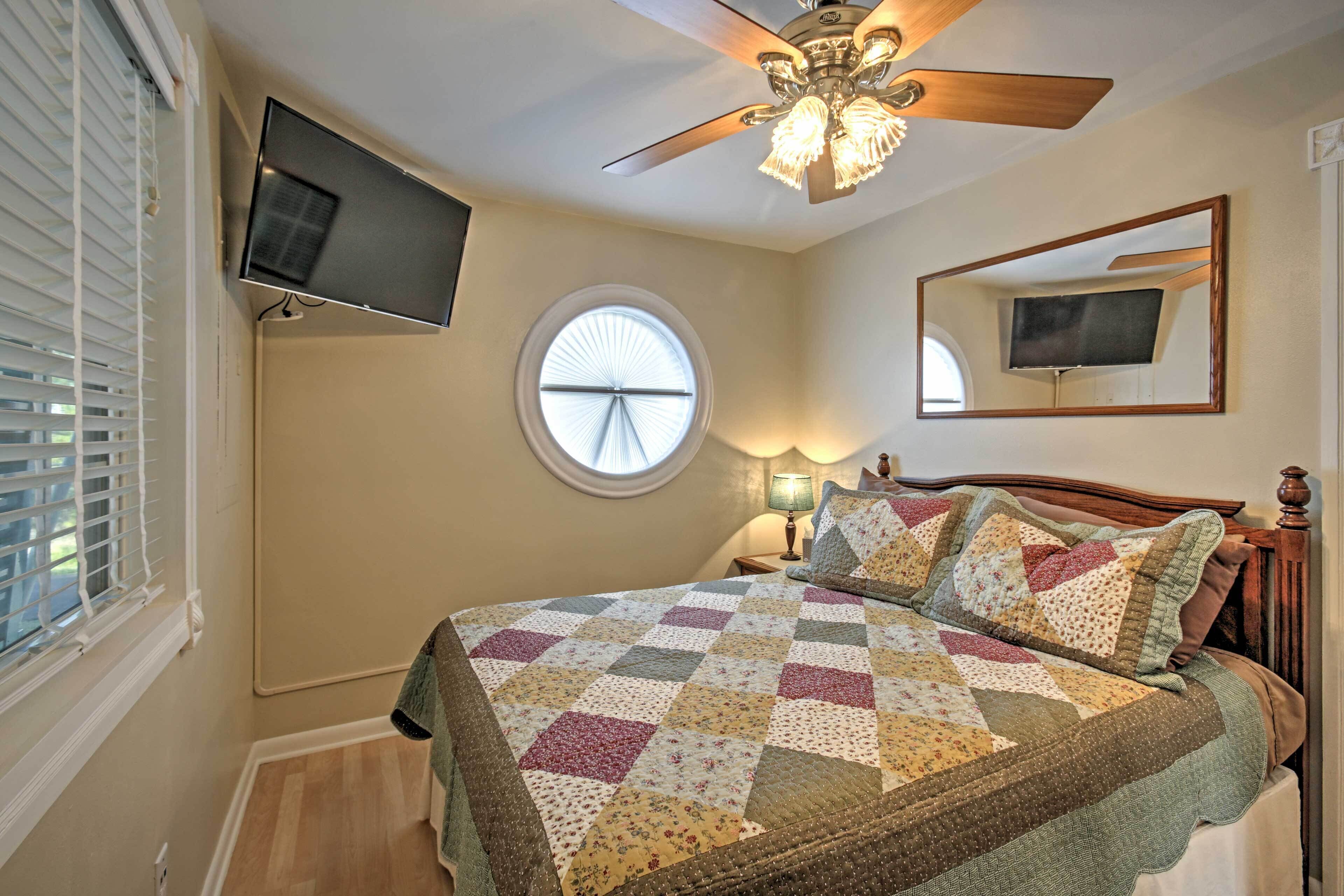 A bedroom with a bed, a ceiling fan, a TV mounted on the wall, a window with blinds, and a round mirror.