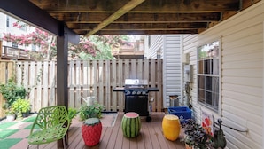 A wooden deck with a barbecue grill, a green chair, a watermelon, and potted plants.