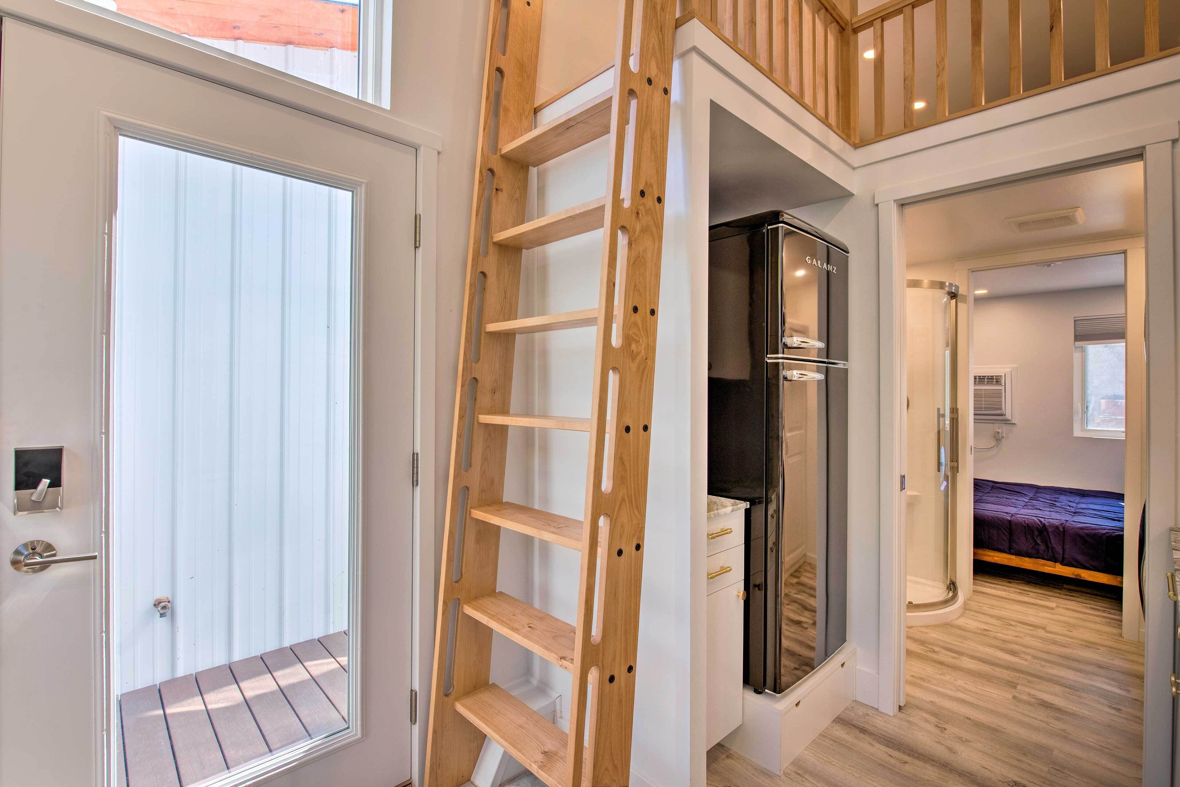 A modern interior with a wooden ladder leading to a balcony, a walk-in closet, and a bedroom with a bed and a window.
