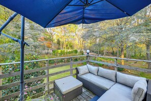 A covered outdoor seating area with a cushioned bench and a table, surrounded by a wooden deck and a garden with trees and shrubs. - Cozy Dix Hills Apt w/ Deck ~ 7 Mi to Beach! (Huntington Station)