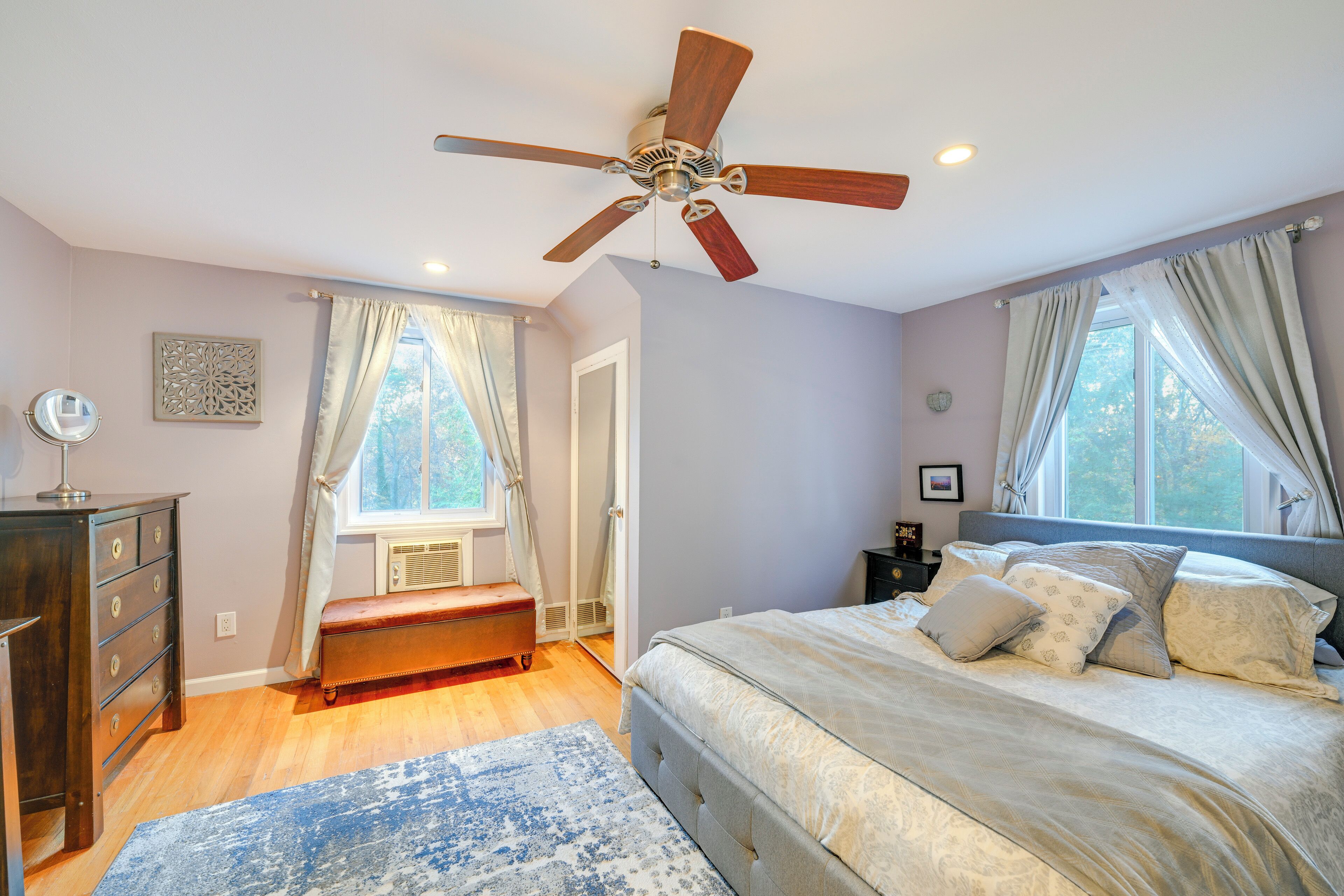 A bedroom with a bed, a ceiling fan, a dresser, a bench, and a window with curtains.