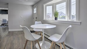 A modern kitchen with white cabinetry, a round white table with four chairs, and a window with a view of houses. - Long Island Home Near Beaches & Golf (Bellmore)