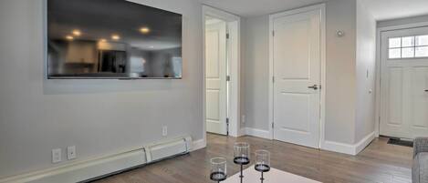 A modern living room with a large flat-screen TV mounted on the wall, a white coffee table with three glasses of red wine, and hardwood flooring.