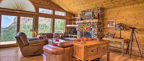 A spacious living room with a wooden ceiling, large windows offering a mountain view, a leather sofa, a wooden coffee table, and a stone fireplace.