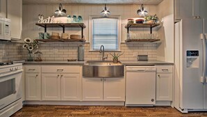 A kitchen with white cabinets, a stainless steel sink, a stove, and a refrigerator. There are two shelves above the sink with jars and dishes.