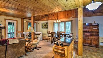 A spacious living room with wooden ceiling and walls, a dining area with a large table and chairs, and a kitchen area with wooden cabinets and a countertop.