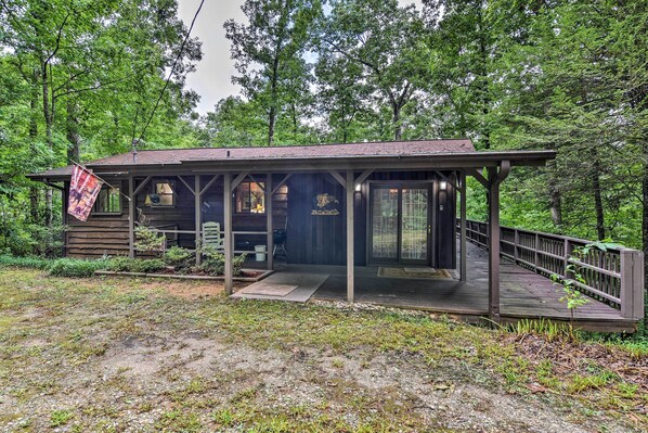 A rustic cabin with a covered porch and a wooden deck, surrounded by dense forest.
