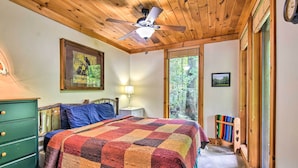 A bedroom with a wooden ceiling, a bed with a colorful quilt, a green dresser, a nightstand with a lamp, and a window with a view of trees. - Robbinsville Cabin w/ Deck: 3 Mi to Fontana Lake! (Robbinsville)