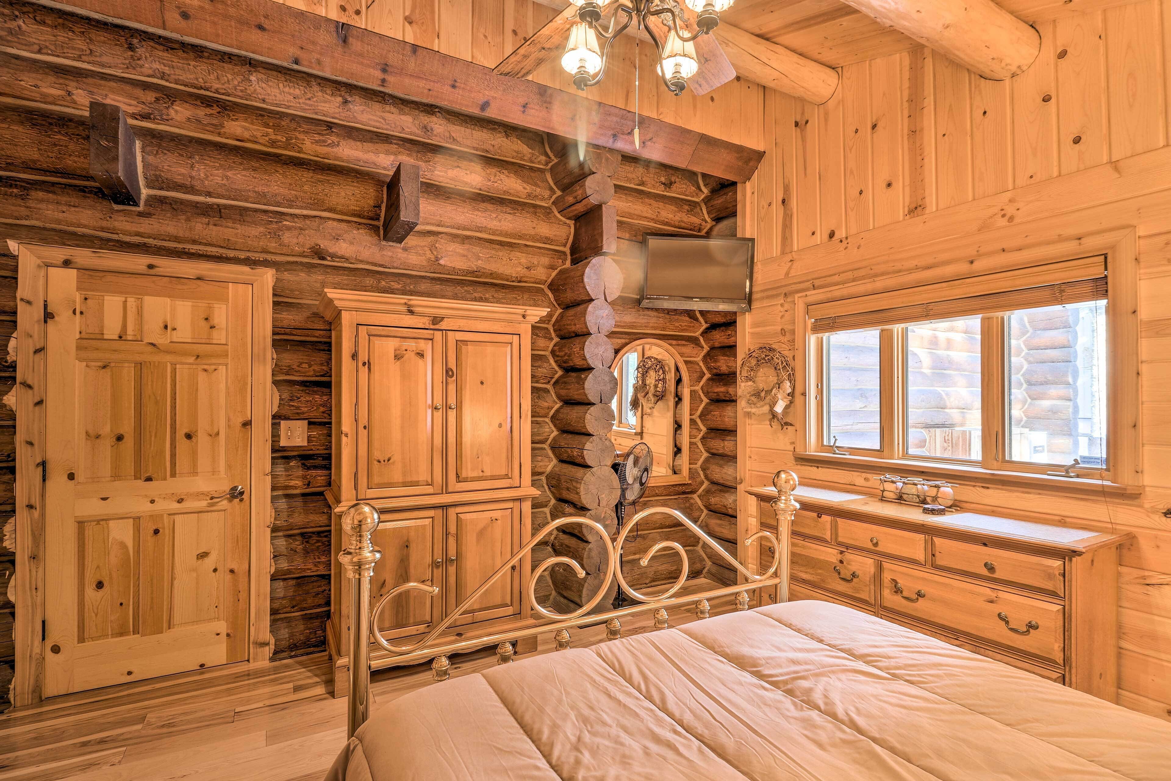 A log cabin bedroom with a bed, wooden cabinets, a dresser, and a stone fireplace.