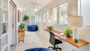 Une pièce moderne avec un bureau en bois, un ordinateur et un fauteuil en forme de poire bleu. On y trouve des fenêtres avec des rideaux, un ventilateur de plafond et une plante en pot sur le bureau.