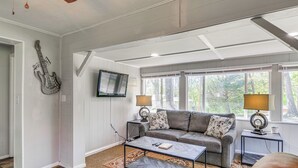 Ein Wohnzimmer mit grauem Sofa, zwei Nachttischen mit Lampen, einem Couchtisch, einem an der Wand befestigten Fernseher und einer Gitarre als Dekoration.