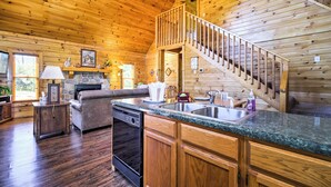 A cozy cabin interior with a wooden interior, a stone fireplace, a kitchen with granite countertops, and a staircase leading to an upper level.