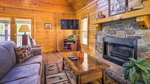 A cozy cabin interior with a stone fireplace, wooden walls, a sofa with a patterned pillow, a coffee table, and a television mounted on the wall.
