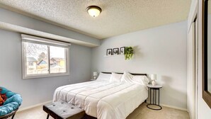 Une chambre à coucher avec un lit, une chaise, une petite table, une fenêtre et un luminaire au plafond.