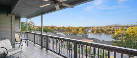 A covered balcony with a ceiling fan, outdoor seating, and a view of a lake and autumn trees.