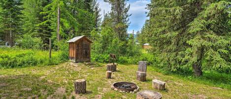 Una cabaña rústica rodeada de un bosque denso, con un pozo de fuego y troncos de madera al frente.