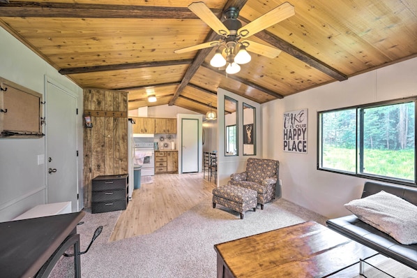 A spacious living area with a wooden ceiling, a ceiling fan, a large window with a forest view, and a cozy seating area.