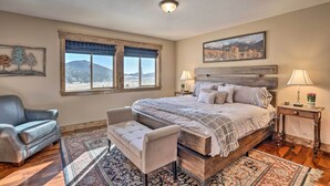 A bedroom with a large bed, a chair, a nightstand, a lamp, and a view of mountains through the window. - Cripple Creek Retreat w/ Incredible Mtn Views! (Cripple Creek)