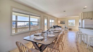 A dining area with a long wooden table set for six, large windows with a view of the beach, and a kitchen area with white bar stools. - Gulf Shores Rental w/ Large Porch - Beach View! (Gulf Shores)
