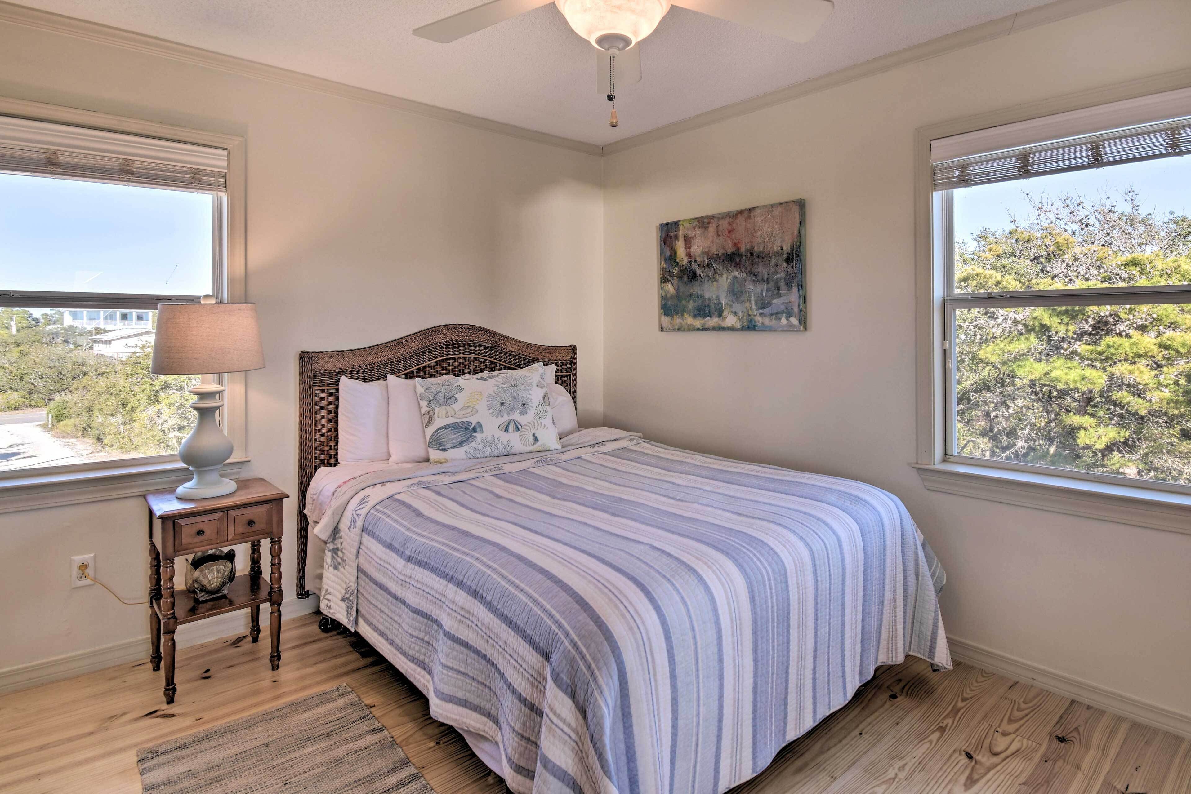 Um quarto com cama, mesa de cabeceira, abajur, quadro e janelas com vista para árvores.