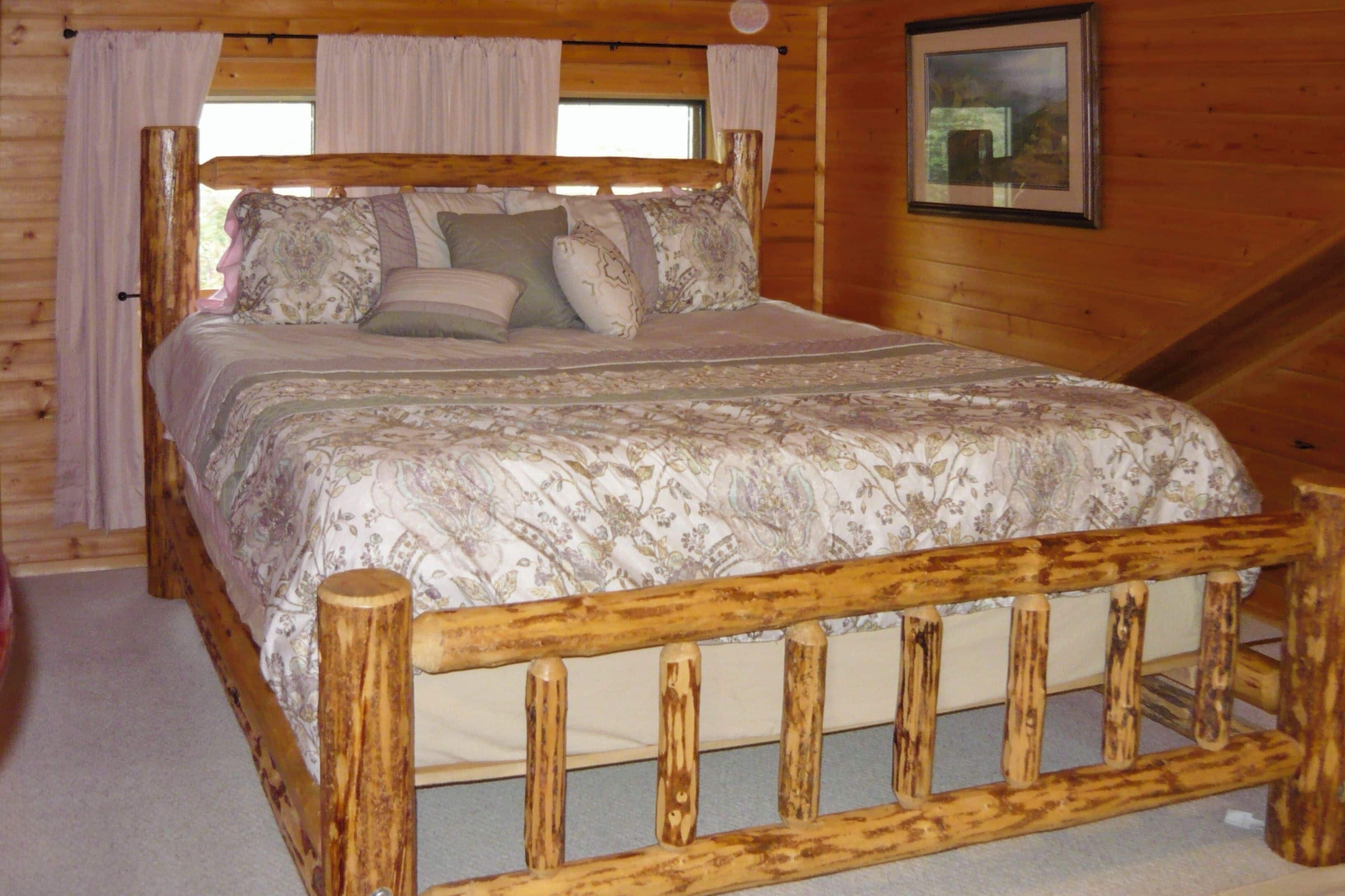 A wooden-framed bed with a patterned bedspread and pillows, a wooden headboard, and a window with curtains.