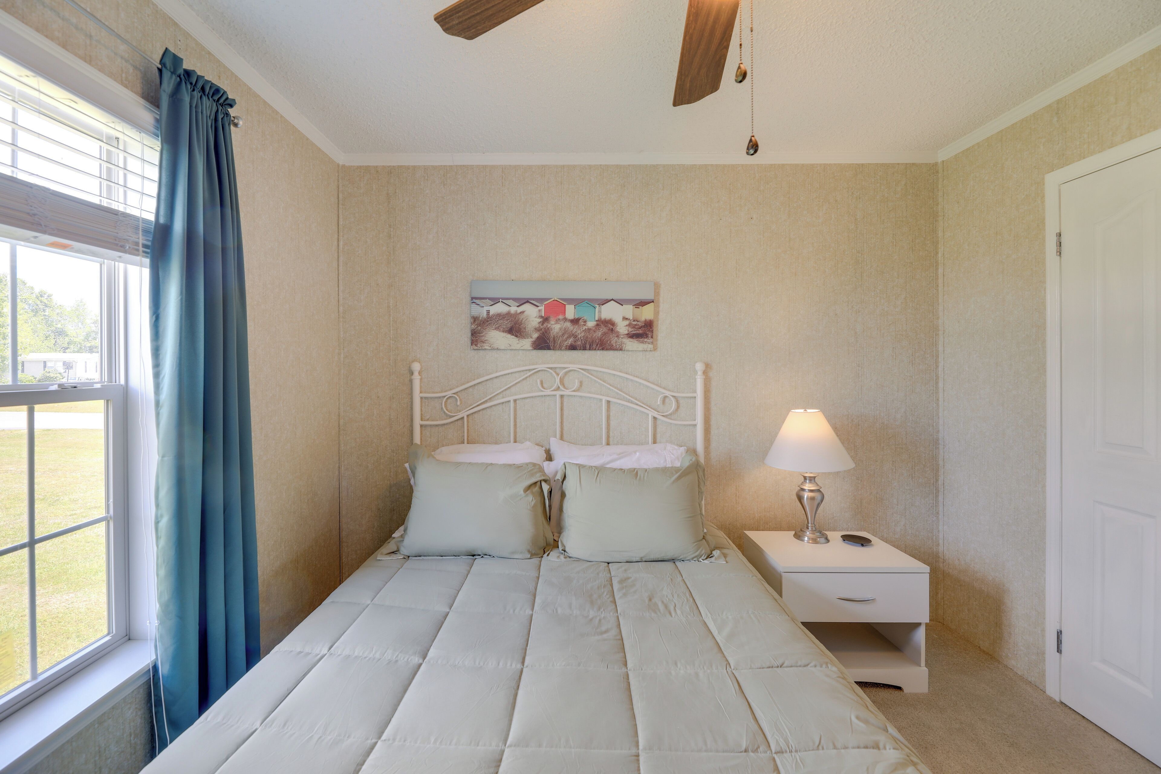 Une chambre à coucher comprenant un lit, une table de chevet, une lampe, un ventilateur de plafond, une fenêtre avec des rideaux et un tableau mural.