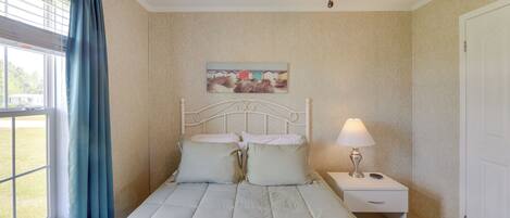 Une chambre à coucher comprenant un lit, une table de chevet, une lampe, un ventilateur de plafond, une fenêtre avec des rideaux et un tableau mural.