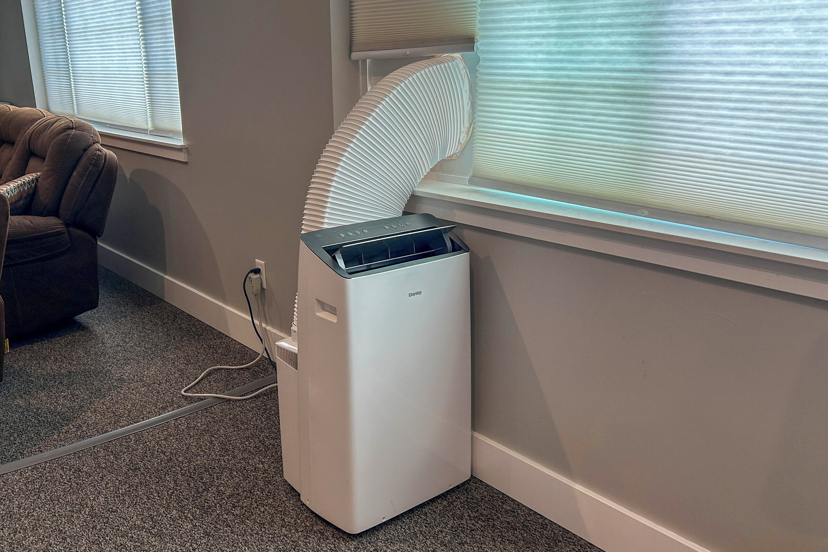 A compact, white portable heater with a control panel, placed against a wall next to a window with blinds.