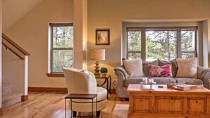 A spacious living room with a high ceiling, a staircase, and large windows offering a view of trees.