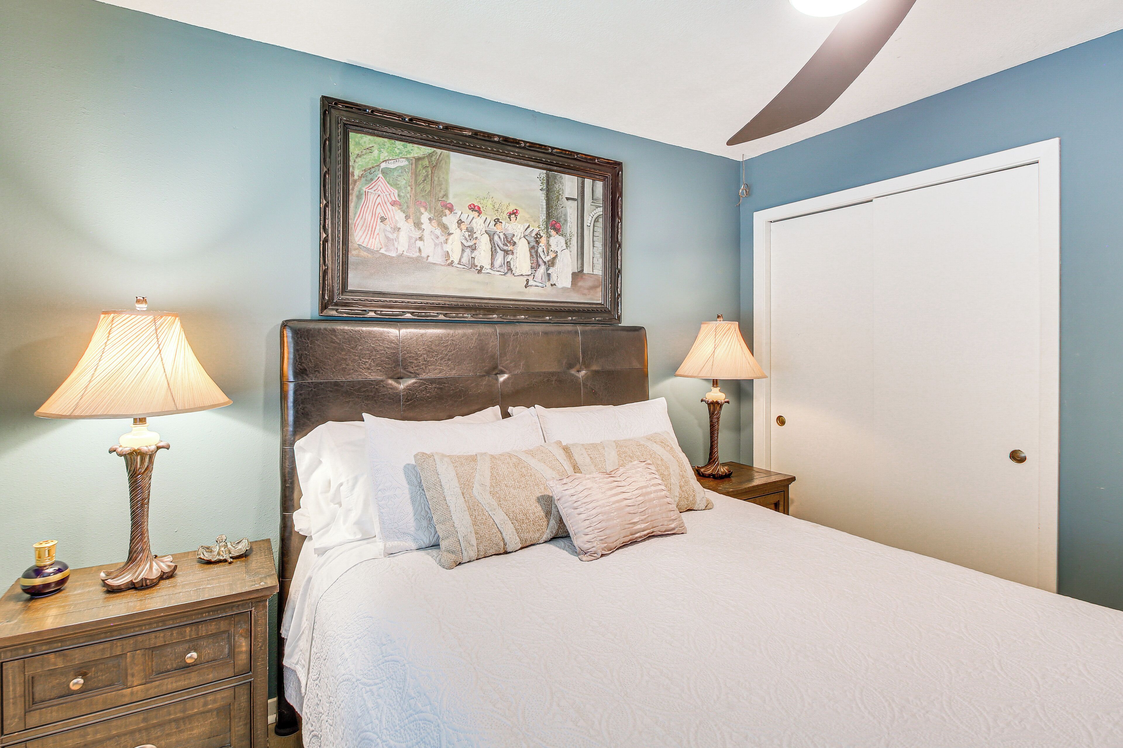 Une chambre à coucher avec un lit, deux tables de chevet, deux lampes, un tableau encadré au mur et une porte.