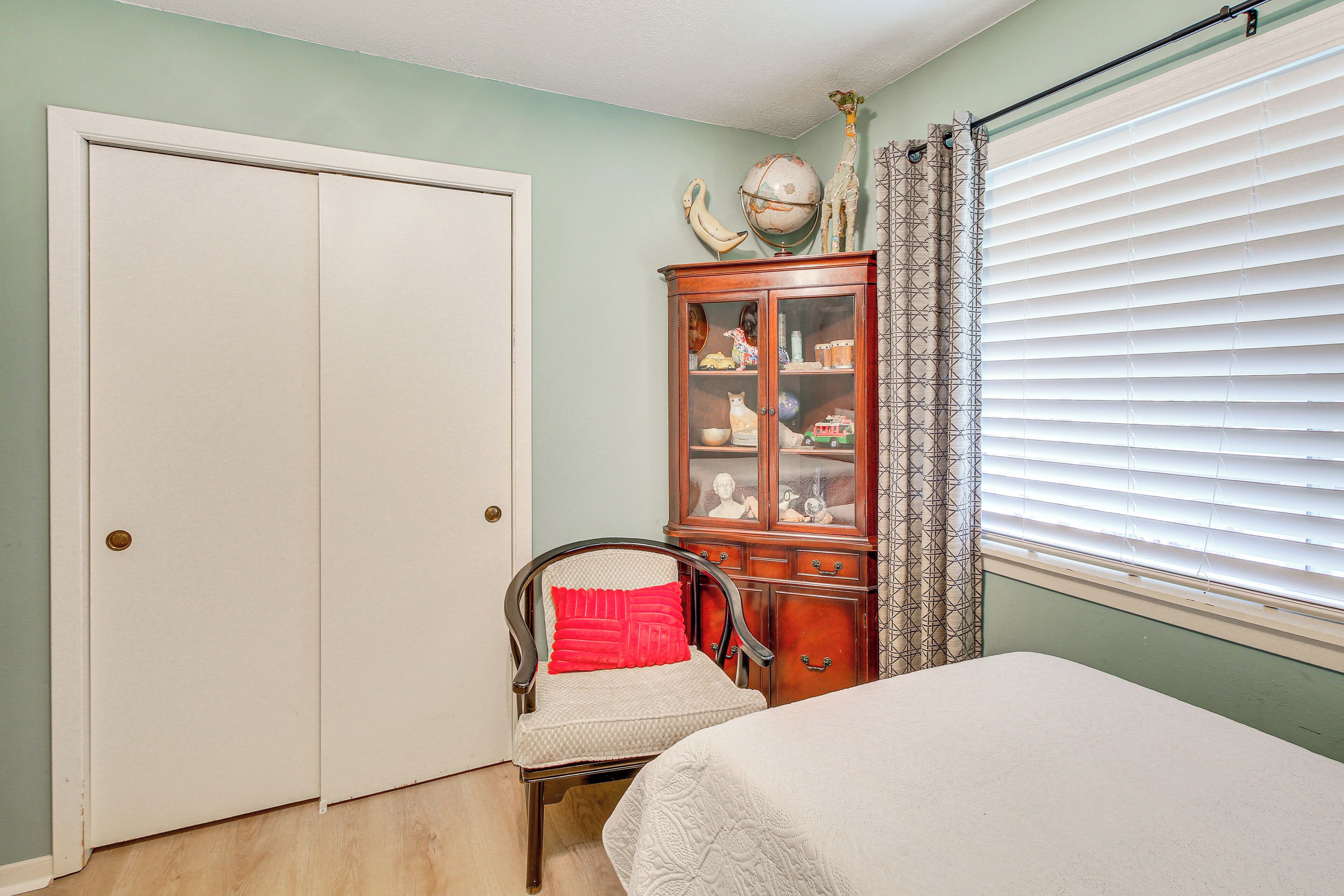 Un dormitorio con una cama, una silla con un cojín rojo, un armario de madera con puertas de cristal y una ventana con persianas.