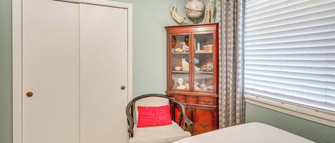 Un dormitorio con una cama, una silla con un cojín rojo, un armario de madera con puertas de cristal y una ventana con persianas.