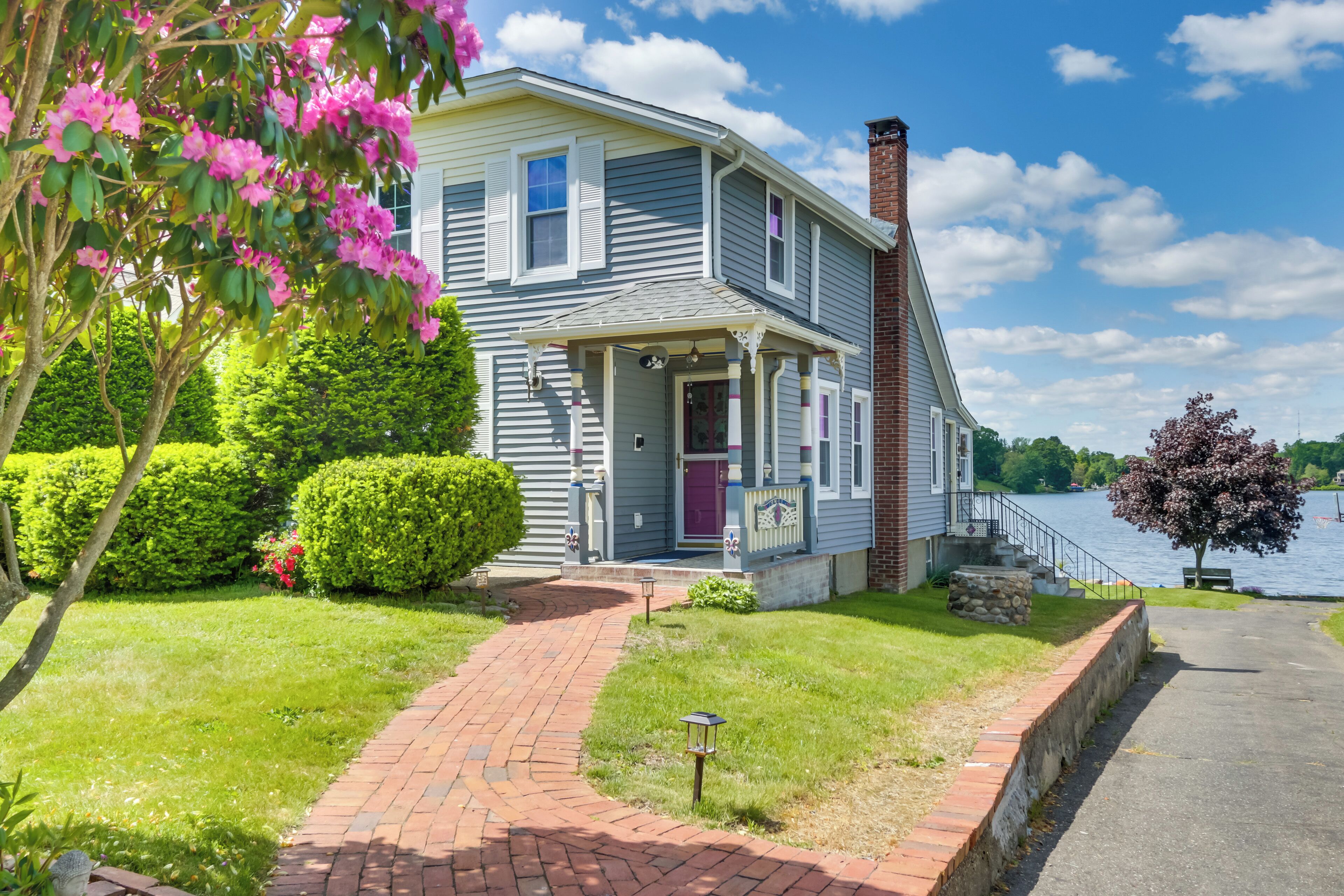 A two-story house with a brick pathway leading to the front door, surrounded by lush greenery and a body of water in the background.