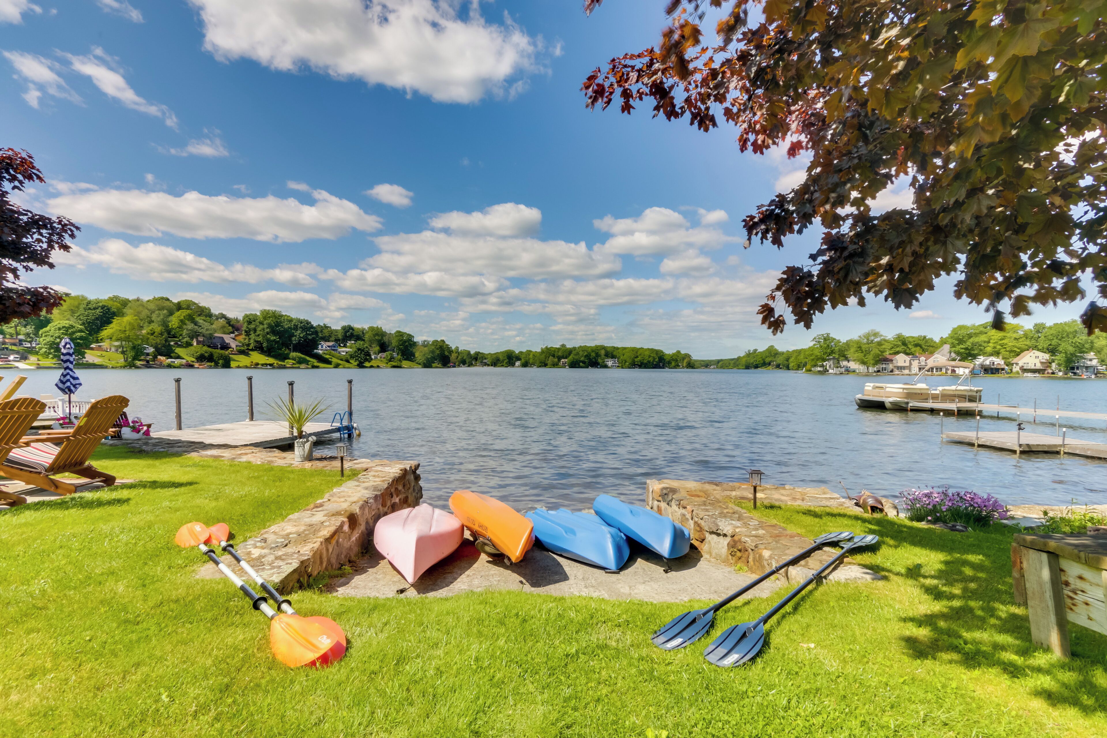 Een landschap aan een meer met kayaks en paddleboards op het gras, een steiger en uitzicht op het water en de omliggende begroeiing.