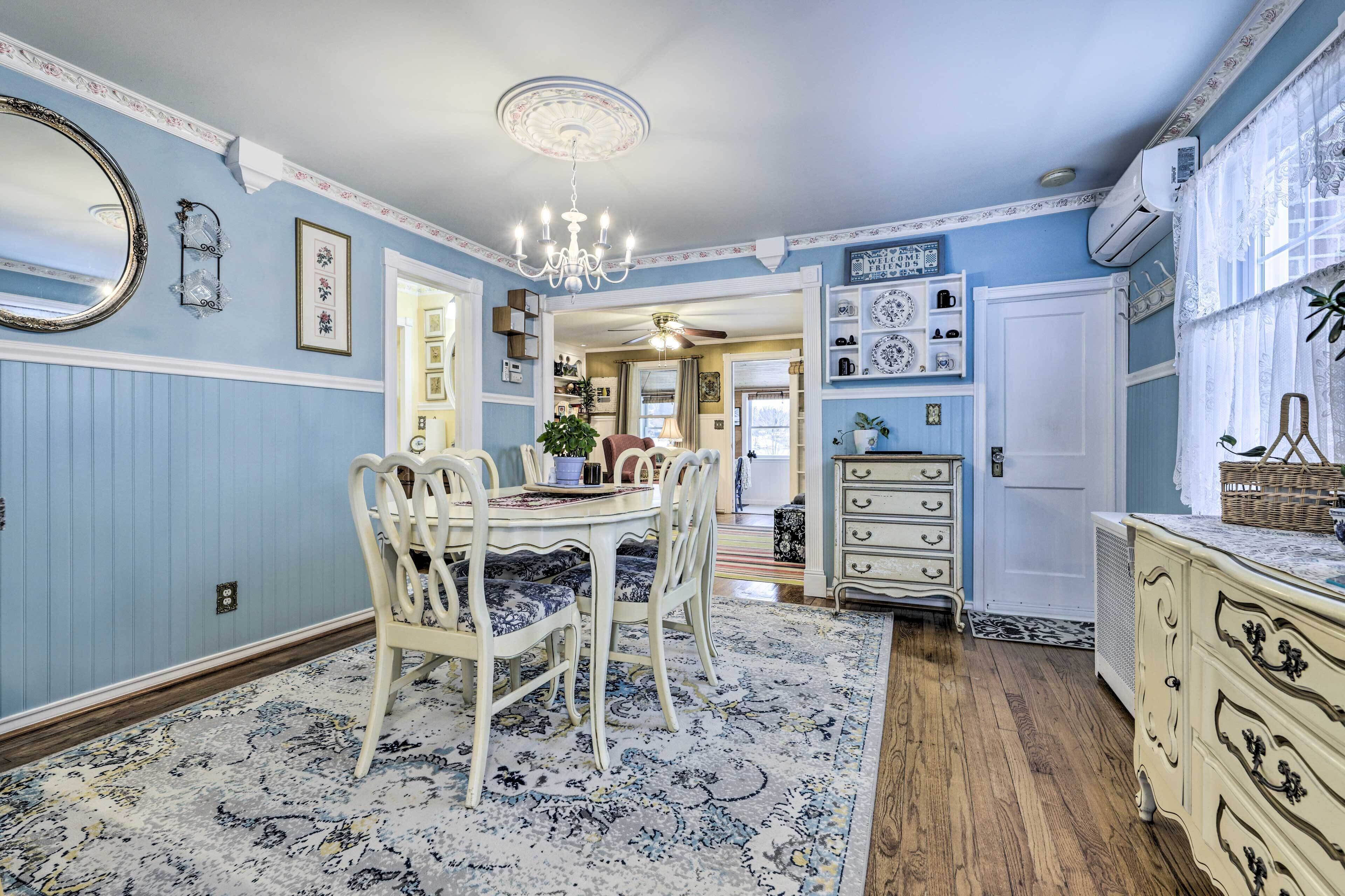 A dining area with a round table, white chairs, and a patterned rug. There is a chandelier above the table and a white dresser with drawers to the right.