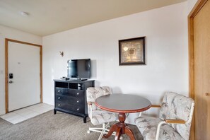 Um quarto com uma mesa de madeira redonda, duas cadeiras estampadas, um aparador preto, uma televisão e um quadro na parede.