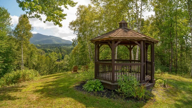 A wooden gazebo in a forested area with a view of mountains in the background.