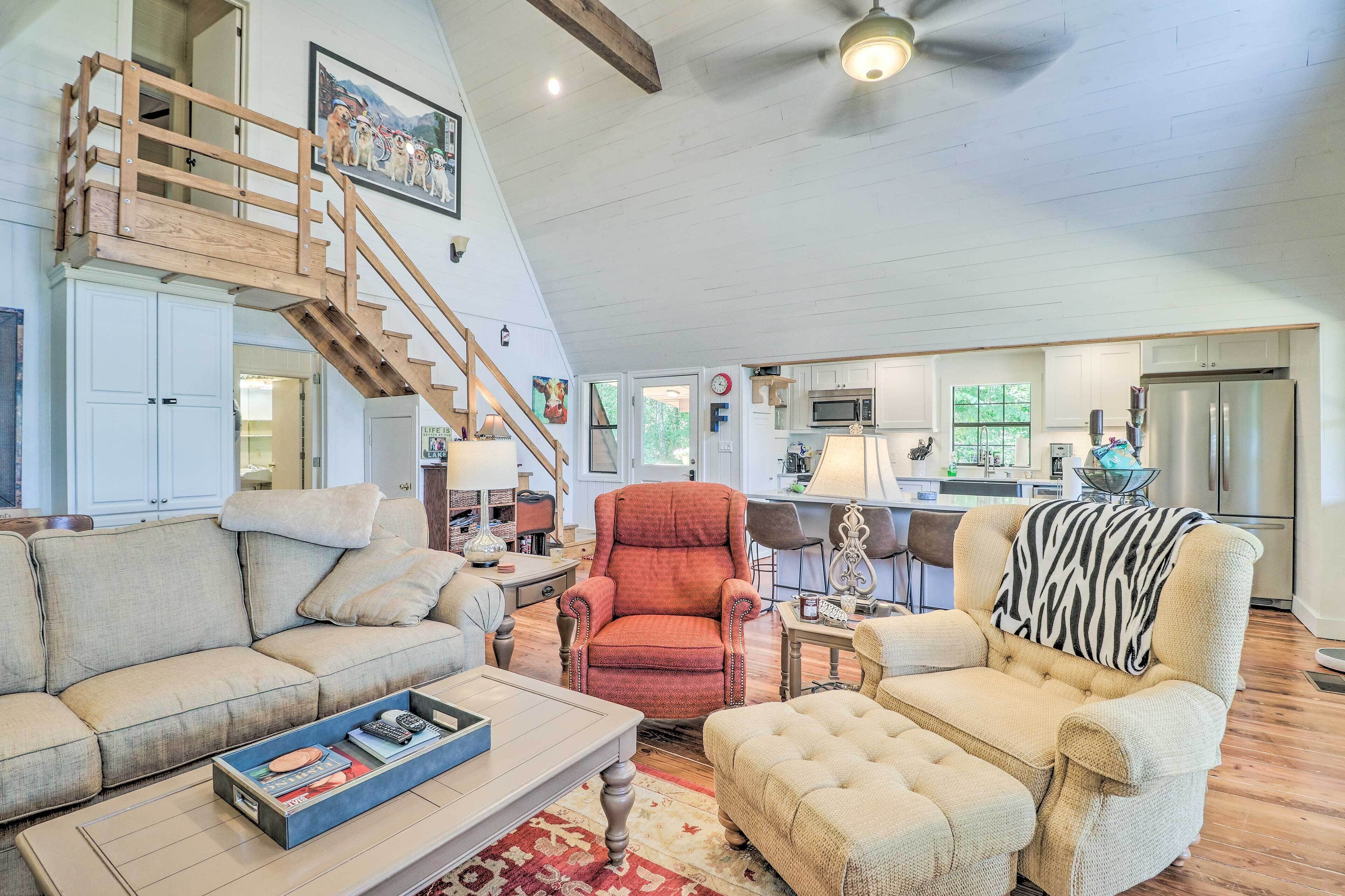 A spacious living room with a high ceiling, a wooden staircase, and a kitchen area visible through an open door.