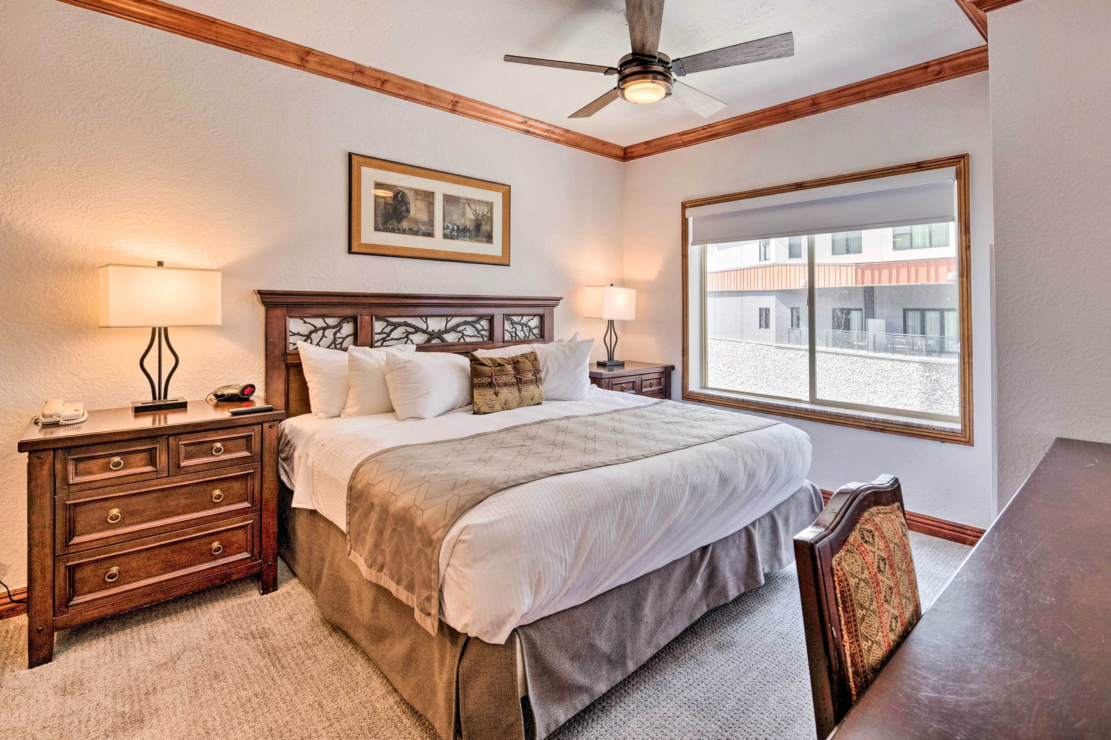Une chambre d’hôtel avec un grand lit, une tête de lit en bois, deux tables de chevet avec des lampes, une fenêtre donnant sur des immeubles et un ventilateur de plafond.