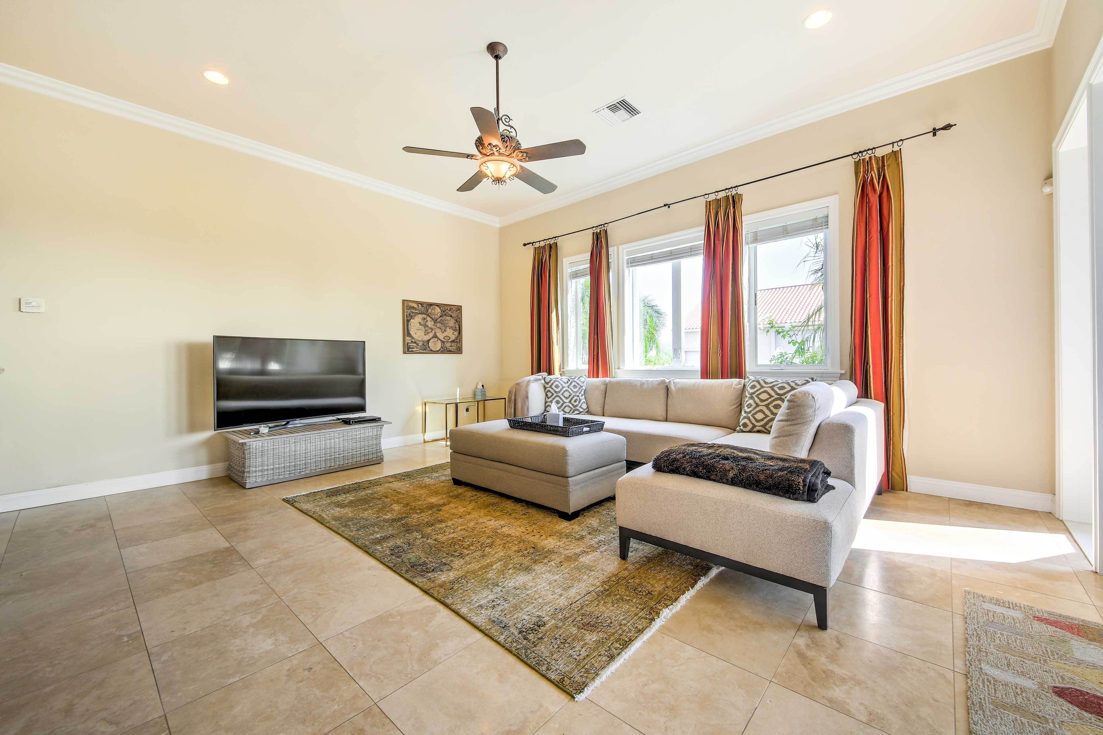A living room with a flat-screen TV, a ceiling fan, a sectional sofa, and a large window with curtains.