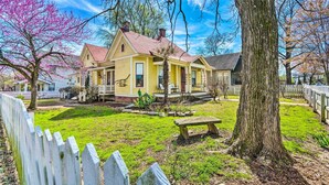 Una casa amarilla con techo rojo, rodeada por una cerca blanca de piquetes, un árbol grande y un banco en un jardín cubierto de césped.