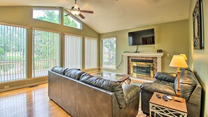 A spacious living room with a leather sofa, a fireplace, a TV mounted on the wall, and large windows.