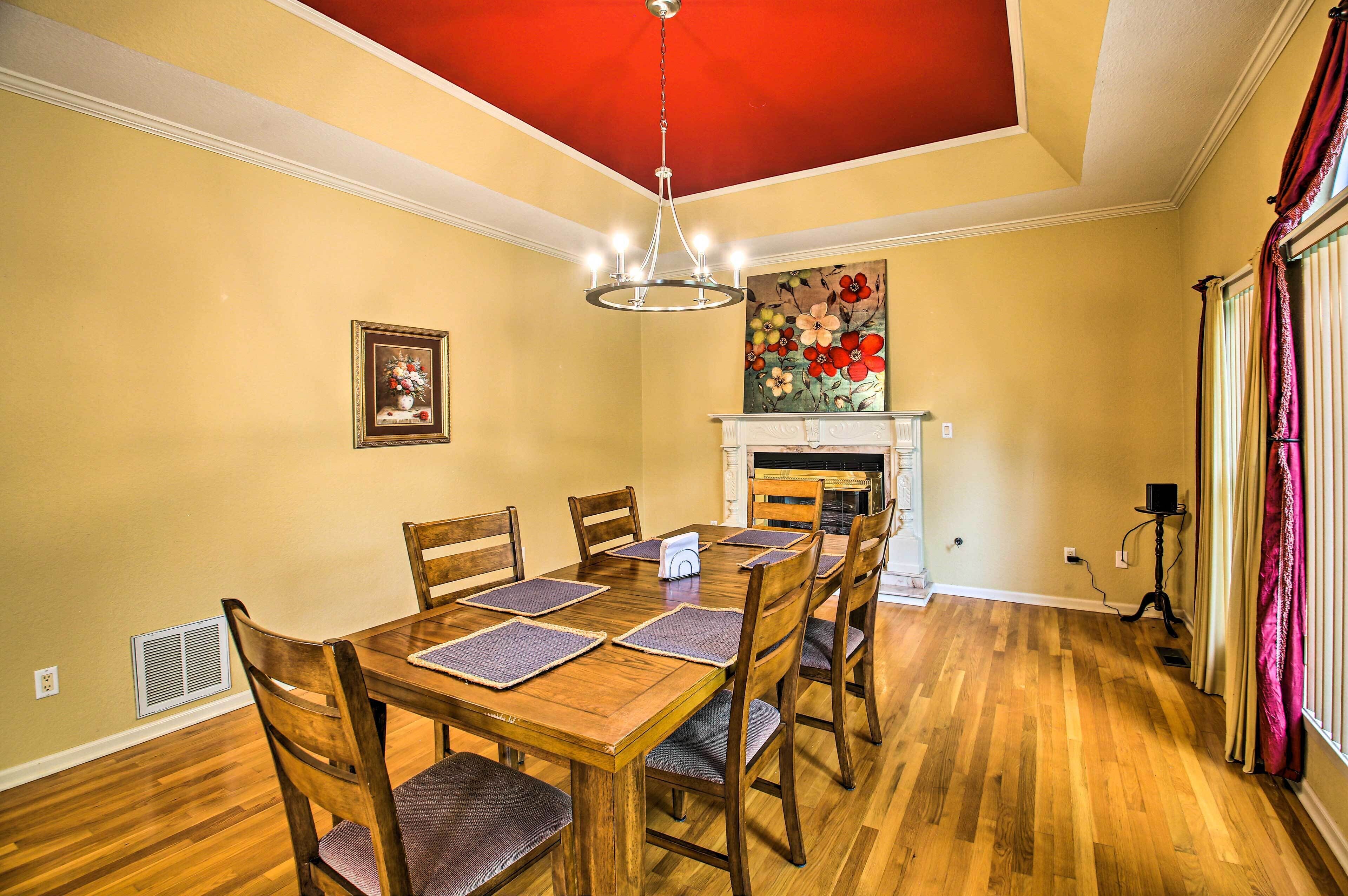 A dining room with a wooden table, chairs, a chandelier, a fireplace, and a painting on the wall.
